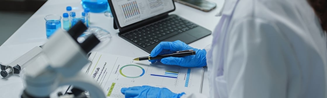 A laboratory worker in protective gloves reviews printed charts beside a microscope, chemical bottles, and an open tablet displaying data.