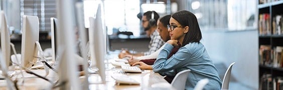 several people seated at a long desk working on desktop computers in what appears to be a library or computer lab. Bookshelves are visible in the background, and the setting is well-lit with natural light coming through large windows