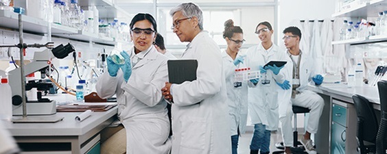 Team of researchers in a laboratory discussing work at a lab bench, with scientific equipment, sample containers, and computer workstations visible throughout the workspace