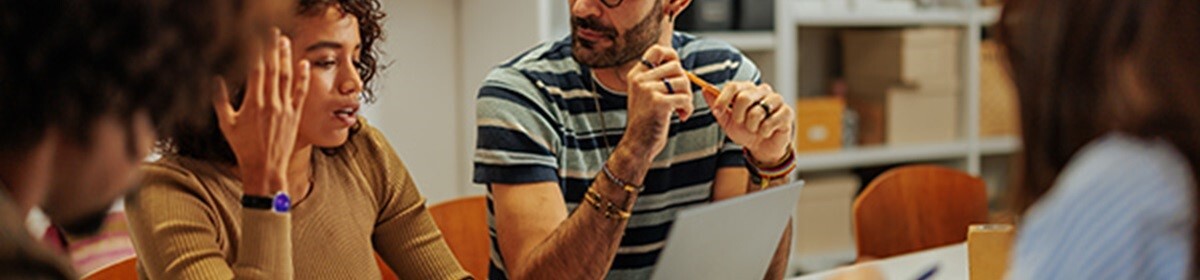 a group of people sitting around a table engaged in a discussion, with one person holding a pencil and another gesturing with their hand