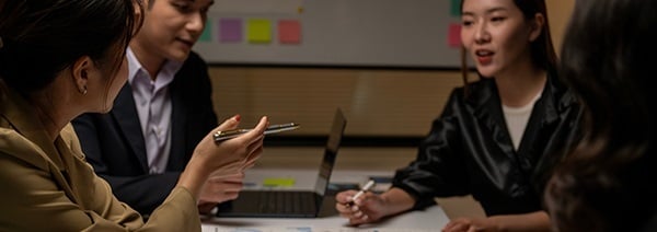 Group of professionals in a meeting, seated around a table with laptops and documents, discussing ideas in front of a whiteboard with colorful sticky notes