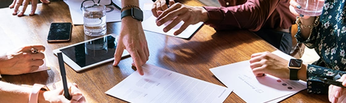Hands of several people gathered around a wooden table reviewing printed documents, with one person pointing at a page while others hold a pen, tablet, and smartphone during a collaborative meeting.
